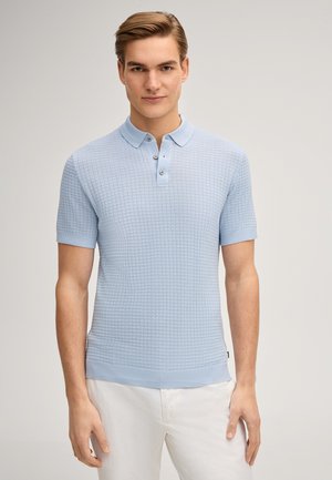 Giovane uomo che indossa una polo azzurra a maniche corte con trama e pantaloni bianchi, in piedi davanti a uno sfondo uniforme.