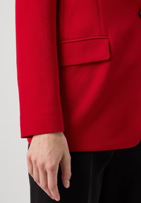 Červený blejzr s hladkým povrchem, zapínání na jeden knoflík a boční kapsou, kombinovaný s černou látkou, ukazující přizpůsobené designové detaily.