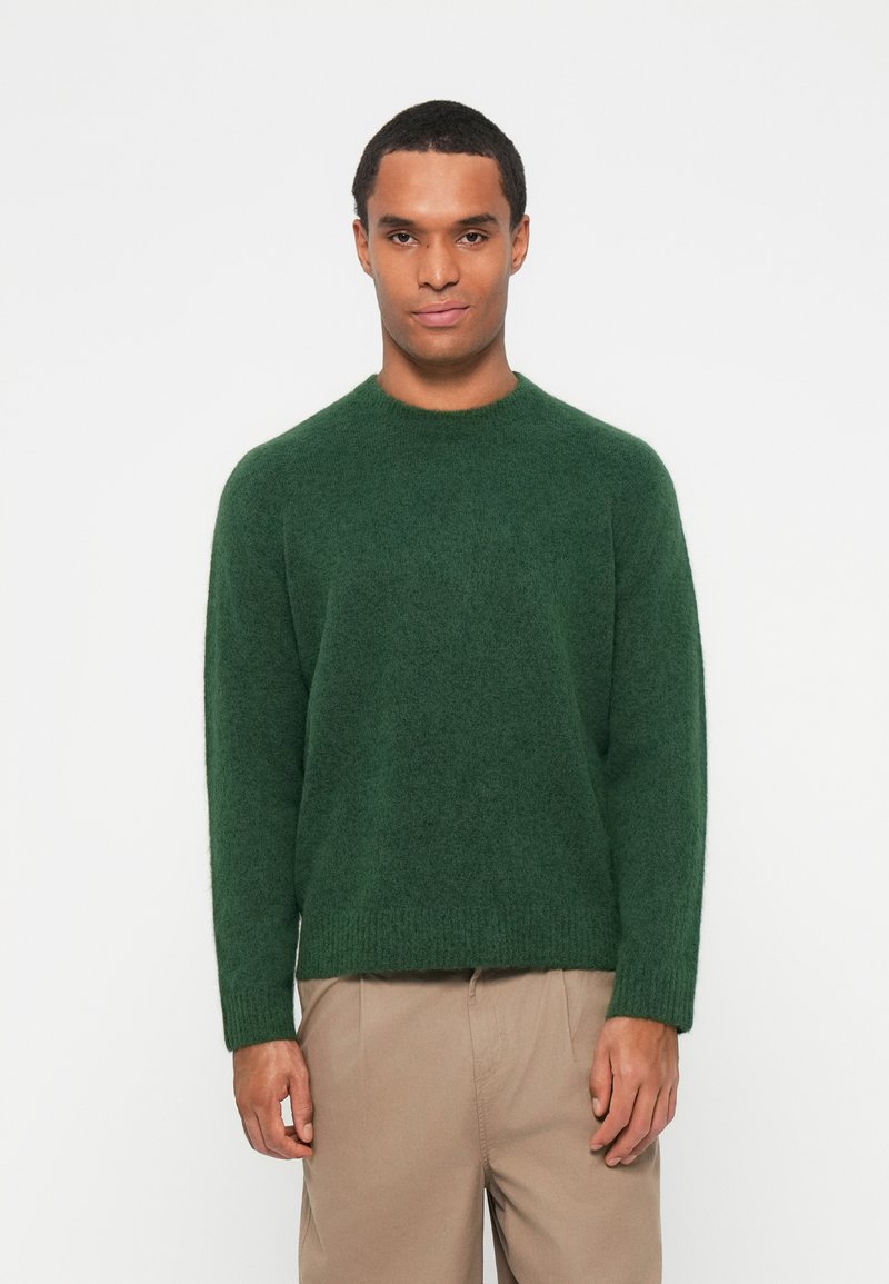 Suéter verde tricotado com mangas longas, gola redonda, textura suave e corte solto, combinado com calças bege.