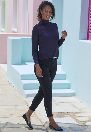 Dunkelviolettes Langarmoberteil, schwarze eng anliegende Hose mit Reißverschlussakzenten und schwarze Ballerinas, vor pastellfarbenen Wänden und Stufen.