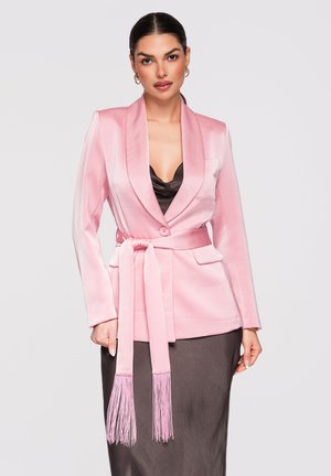 Vrouw draagt een roze satijnen blazer met ceintuur over een zwarte satijnen jurk, met gouden oorringen en strak naar achteren getrokken haar.