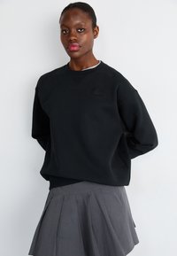 Sudadera negra de corte relajado, con escote redondo y un pequeño logo en el pecho, combinada con una falda gris plisada.