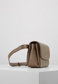 Sac à main en similicuir beige avec une forme structurée, un rabat pliant et une bandoulière réglable. Présente un logo doré discret et une surface texturée.