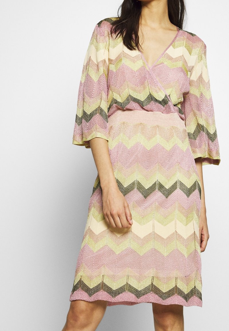 Femme portant une robe jusqu'aux genoux avec des motifs en zigzag rose, jaune, crème et noir, et des manches larges arrivant aux coudes.