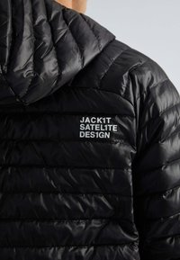Sort polstret jakke med hætte. Har en quilted tekstur og trykt logo "JACK1T SATELITE DES1GN" i hvid.