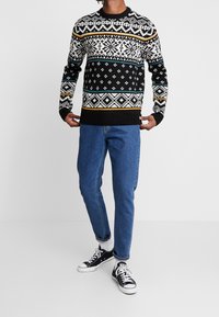 Pull en tricot noir avec des motifs géométriques blancs et colorés, col rond, poignets et ourlet côtelés, associé à un jean bleu et des baskets noires.