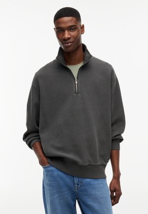 Ung mand iført en mørkegrå kvart-lynlås trøje og blå jeans, stående med den ene hånd i lommen mod en ensfarvet hvid baggrund.