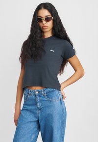 Svart kortermet t-skjorte med hvit "obey"-tekst, parret med løse blå denimjeans. Brukt med grønntinters cat-eye solbriller.