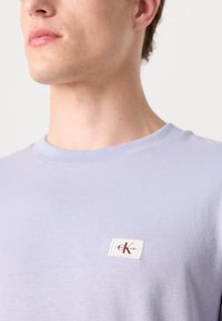 Világoslila, kerek nyakú póló, puha pamutból készült, melyen egy kicsi, fehér és piros logófolt található a mellrészen. Simább textúra, minimális dizájn.