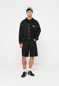 Black denim jacket with two chest pockets, paired with a black t-shirt and black knee-length shorts. Black sneakers with white accents.