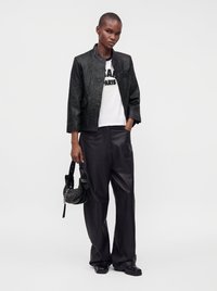 Woman standing wearing black leather jacket, white graphic t-shirt, black wide-leg pants, holding a small black handbag with silver studs.