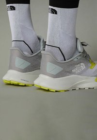 Graue Sportschuhe mit einem Obermaterial aus Mesh, weißer Sohle und gelben Akzenten. Dazu weiße Socken mit einem schwarzen Logo. Sichtbare designelemente mit Textur.