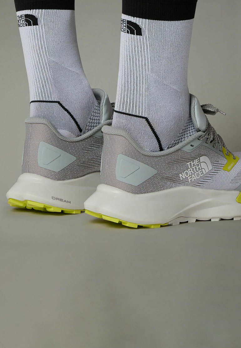 Graue Sportschuhe mit einem Obermaterial aus Mesh, weißer Sohle und gelben Akzenten. Dazu weiße Socken mit einem schwarzen Logo. Sichtbare designelemente mit Textur.
