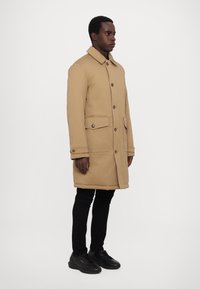 Beige wool blend coat with a button front, collar, two front pockets, and a longer silhouette. Paired with black slim-fit trousers and sneakers.