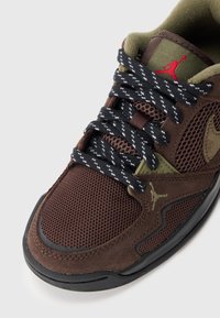 Brune joggesko med mesh- og semsket skinnmaterialer, svarte og hvite mønstrede snører, grønne detaljer og en rød logo på tungen.