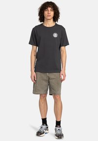 Donkergrijze katoenen t-shirt met een logo op de voorkant, gecombineerd met kaki shorts. Model draagt zwarte sokken en meerkleurige sneakers, staand voor een witte achtergrond.