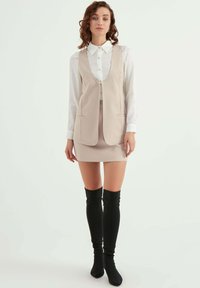 Beige ärmlös väst över en vit skjorta med knappar, i kombination med en figurnära beige kjol och svarta över-knä-höga stövlar. Slät tygtextur.