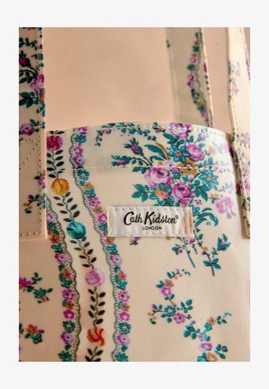 Gėlėmis raštuotas audeklo krepšys kreminės spalvos, su rožinėmis ir mėlynomis gėlėmis, žaliomis lapėmis bei siūtais etiketėmis su užrašu "Cath Kidston, London."