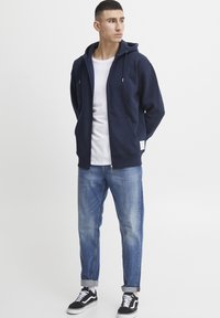Navy zip-up hoodie with a drawstring hood, paired with a white t-shirt, light blue denim jeans, and black sneakers with white accents.