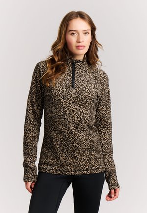 Ung kvinde med bølget hår iført en leopardmønstret langærmet top med lynlås foran, stående mod en ensfarvet lys baggrund.