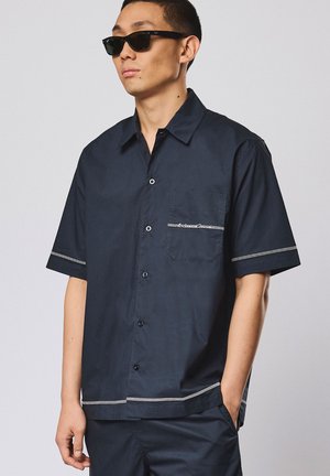 Mann trägt schwarze Sonnenbrille und ein dunkelblaues kurzärmliges Hemd mit weißen Rändern, steht mit einer Hand in der Tasche.