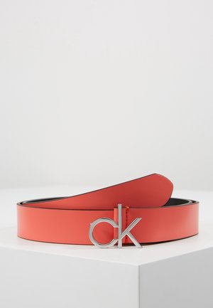 Rødt læderbælte med sølv "ck" logo spænde, som hviler på en hvid overflade mod en ensartet lys baggrund.