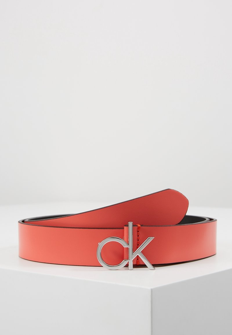Ceinture en cuir rouge avec boucle en argent au logo "ck", posée sur une surface blanche sur fond clair uni.