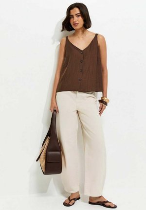 Femme portant un haut marron sans manches boutonné, un pantalon crème à jambes larges, des sandales, un sac à main marron avec un détail tressé, et des bracelets.