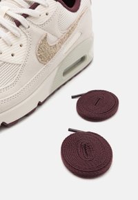 Nike Air Max kedas ir ar krēma krāsas augšdaļu, kurai ir teksturēts bordo Swoosh, kombinēti ar divām apaļām bordo auklām uz baltā fona.