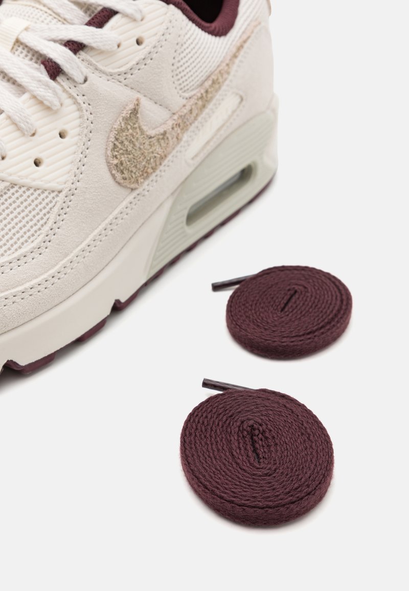 De Nike Air Max sneaker heeft een crèmekleurig bovenwerk met een gemarmerde bordeauxrode Swoosh, gecombineerd met twee ronde bordeauxrode veters op een witte achtergrond.