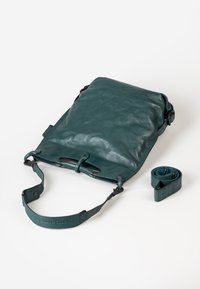 Groene leren tas met een gladde textuur, een opvouwbaar ontwerp, een verstelbare schouderband en een opgerolde riemaccessoire ernaast geplaatst.