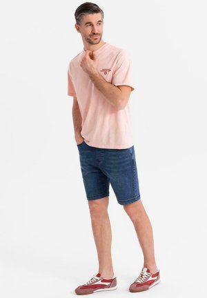 Lys pink bomulds t-shirt med korte ærmer og et lille brystlogo, parret med blå denim shorts og røde og hvide sneakers.