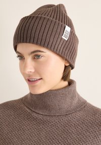 Bonnet en tricot côtelé marron avec un revers et une étiquette blanche ornée d'un motif de vague. La personne porte un pull à col montant assorti.