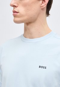 Νεαρός άνδρας που φοράει γαλάζιο μπλουζάκι με στρογγυλή λαιμόκοψη και μαύρο λογότυπο "BOSS" στο στήθος, εμφανιζόμενος από τον λαιμό μέχρι το άνω μέρος του κορμού σε ουδέτερο φόντο.