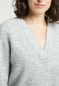 Pull en maille gris clair avec un design en V, présentant une surface texturée et des détails côtelés le long de l'encolure et des manches.