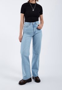 Personne portant un haut noir à manches courtes, un jean droit taille haute bleu clair, des colliers en argent superposés et des chaussures compensées marron foncé.