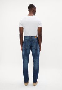 Blugi închisi la culoare, cu croială dreaptă, având un eticheta pe brâu la spate, cinci buzunare și o decolorare subtilă. Combinați cu un tricou alb.