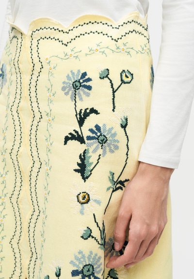 Χέρι που ακουμπά σε κίτρινη φούστα με κεντημένα μπλε και λευκά λουλούδια, πράσινα φύλλα και μαύρη ραφή με κυματιστό σχέδιο.