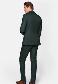 Ciemnozielony garnitur o fakturowanym wykończeniu, z dopasowaną marynarką i zwężającymi się spodniami. Brązowe skórzane buty dopełniają całość.