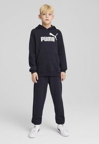 Tumši zils kapucis ar baltu "PUMA" logo, ar priekšējo kabatu un ribotām aprocēm, kopā ar atbilstošiem tumši ziliem sporta biksēm un baltām kedām.