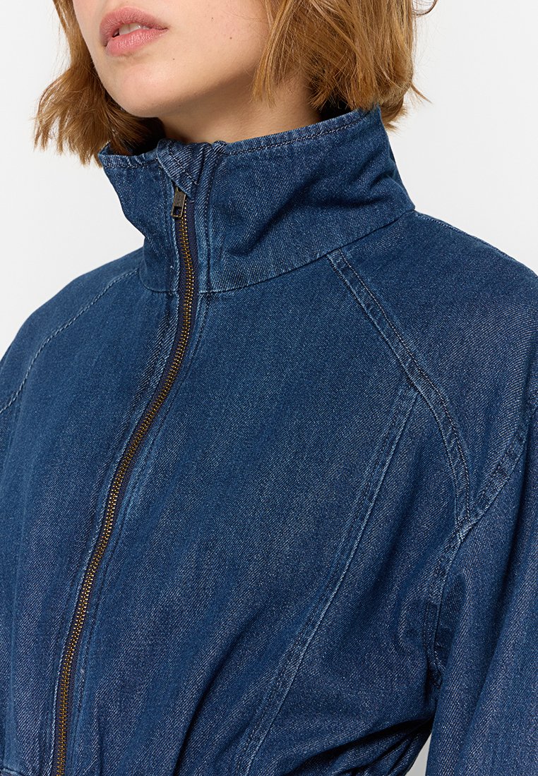 La chaqueta de jeans presenta un cuello alto, cremallera completa y mangas raglán. El material es de color azul oscuro con detalles de costura visibles.