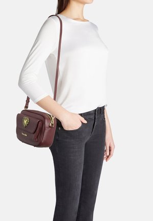 Borsa a tracolla in pelle bordeaux con hardware dorato, dettaglio a forma di cuore sul davanti e una lunga tracolla, posizionata su uno sfondo bianco.