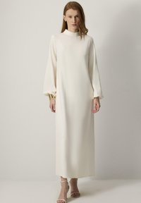 Lange, crèmekleurige jurk met een hoge halslijn en lange, volumineuze mouwen. Bevat gouden accenten bij de manchetten en heeft een gladde textuur.