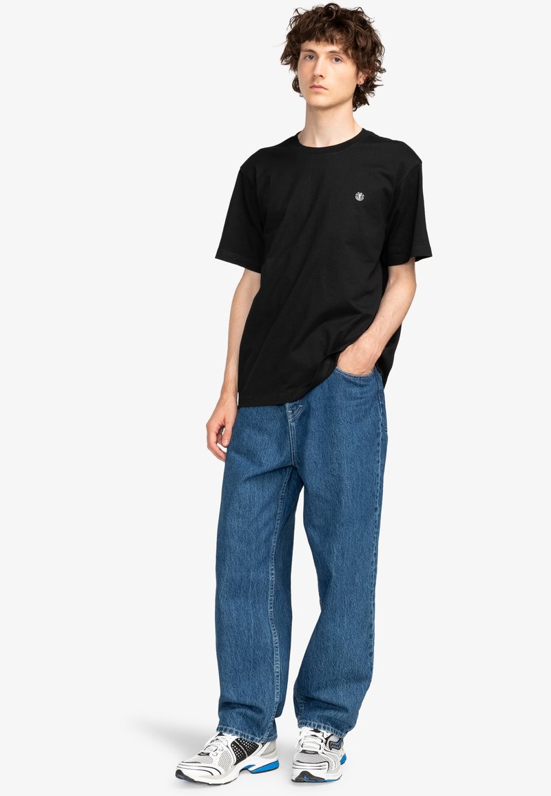 Czarna koszulka z krótkim rękawem z małym logo, zestawiona z luźnymi niebieskimi dżinsami i białymi trampkami z czarnymi akcentami.