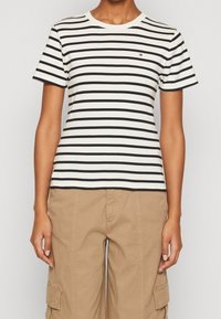 Pruhované tričko s krátkým rukávem v bílé a černé barvě, s kulatým výstřihem a malým logem na hrudi. Materiál je hladký a přiléhavý.