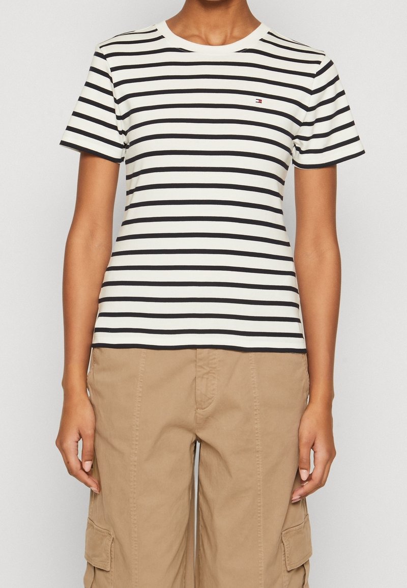 Pruhované tričko s krátkým rukávem v bílé a černé barvě, s kulatým výstřihem a malým logem na hrudi. Materiál je hladký a přiléhavý.