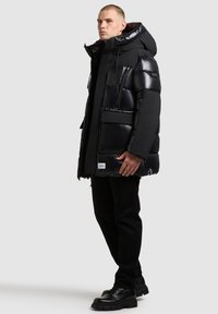 Must must jacket, mille kaelaga, kapuutsi ja mitme taskuga. Läikiv viimistlus, paks vooder ja ruuduline mustri detail.