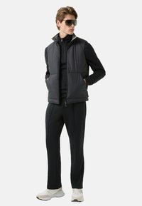 Czarna pikowana kamizelka na czarnej bluzie z zamkiem, w połączeniu z czarnymi spodniami i białymi trampkami. Modelka nosi duże okulary przeciwsłoneczne.