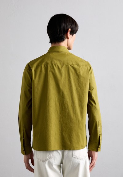 Olivno zelena srajca z dolgimi rokavi, s ovratnikom in manšetami na gumb, izdelana iz teksturiranega materiala; nošena z svetlimi hlačami.