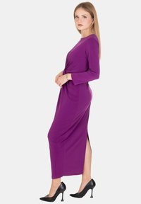Abito lungo viola con maniche lunghe, spacco laterale e dettaglio a torsione in vita, abbinato a scarpe nere con tacco alto. Tessuto liscio.